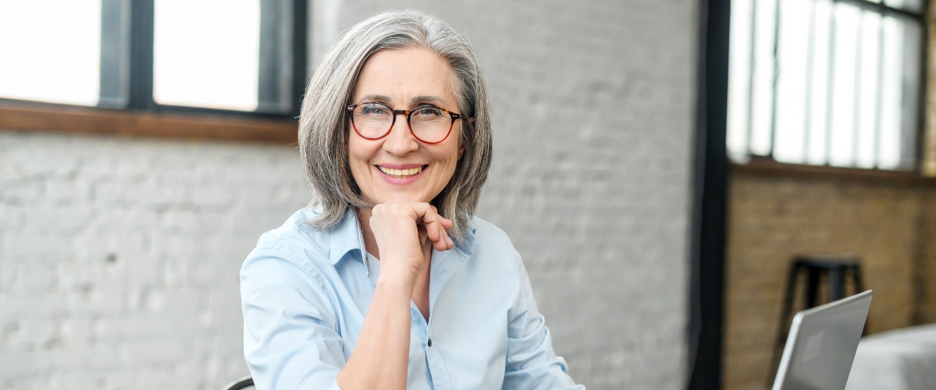 Intelligent elderly gray-haired businesswoman using a laptop in the office. Smart mature woman entrepreneur looks at the camera sitting at the desk. Senior online teacher, elderly female employee